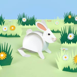 May include: A white paper rabbit with pink-lined ears and a small tail, positioned in a papercraft garden scene. The scene includes green grass, white and yellow paper flowers, and a blue sky background. The rabbit is the focal point.