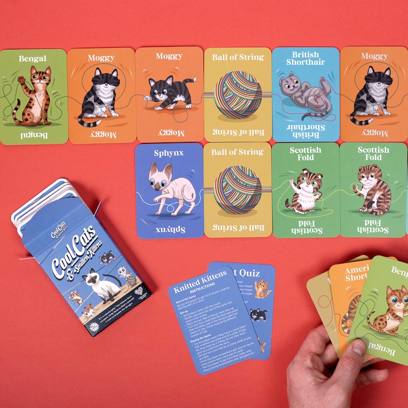 May include: A card game set featuring illustrated cat breeds and a ball of string. The game box reads "Cool Cats & Smitten Kittens." Additional cards include a quiz and instructions. The cards are arranged on a red surface.