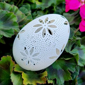 May include: A white, egg-shaped decorative object with a floral cut-out design. The object is surrounded by green leaves and pink flowers, creating a natural backdrop. The egg-shaped object is approximately 5 inches in length.