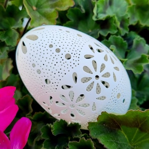 May include: A white, egg-shaped decorative object with intricate floral cutouts and small dot perforations. The object is set against a backdrop of green foliage and a vibrant pink flower, creating a contrast of textures and colors.