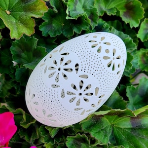May include: A white, egg-shaped decorative object with a floral cut-out design. The object is set against a backdrop of green leaves and a pink flower. The egg-shaped object is likely a candle holder or decorative piece.