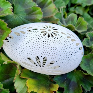 May include: A white, oval-shaped decorative object with intricate floral cutouts and small perforations. The object is set against a backdrop of green leaves, creating a natural contrast. The design suggests a decorative home accent.
