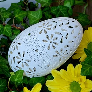 May include: A white, oval-shaped decorative object with a floral cutout design. The object is surrounded by green ivy and bright yellow flowers, creating a natural and decorative display. The object is likely ceramic or porcelain.