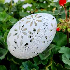 May include: White, egg-shaped decorative object with floral cutouts and small perforations. The object is set against a backdrop of green leaves and red flower buds, creating a natural contrast. The design is intricate and delicate.