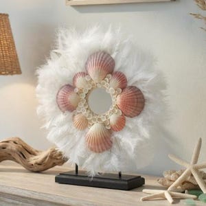 May include: A seaside-inspired display with a woven lamp, driftwood, and a seashell wreath. A blue glass bottle vase holds dried grasses. A framed watercolour painting of a seascape hangs on the wall.