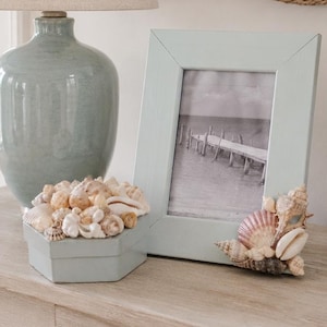 May include: A light blue picture frame decorated with various seashells on the bottom right corner, positioned next to a matching light blue hexagonal box filled with seashells, a light blue ceramic lamp, and a white textured vase with greenery.