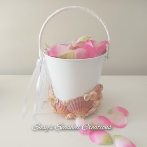 May include: White metal bucket with a white handle, filled with pink and green rose petals. The base is decorated with seashells. White ribbons are tied to the side. The text "Susy's Seashell Creations" is at the bottom.
