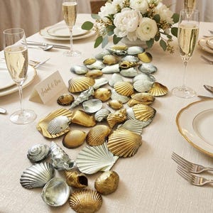 May include: A table centrepiece arrangement consisting of numerous assorted seashells painted in metallic gold and silver finishes, scattered across a light-coloured tablecloth between champagne flutes, white plates, and a floral bouquet.