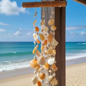 Könnte beinhalten: Ein Muschel-Windspiel hängt an einem Holzbalken, mit einem Strand und Ozean im Hintergrund. Das Windspiel besteht aus verschiedenen Muscheln in Weiß-, Orange- und Cremetönen, die mit weißer Schnur aufgefädelt sind. Der Strand hat goldenen Sand und der Ozean ist leuchtend blau.