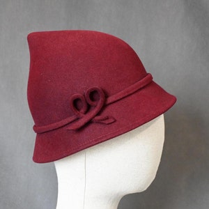May include: A claret-coloured cloche hat with a rounded crown and a small, downward-sloping brim. The hat has a decorative bow on the front and a band around the crown. The hat is made of a felt-like material.