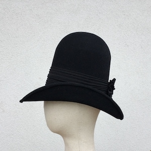 Może przedstawiać: Czarny kapelusz cloche z zaokrągloną koroną i szerokim rondem. Kapelusz posiada plisany pasek wokół korony i mały, dekoracyjny kwiat z boku. Kapelusz jest prezentowany na głowie manekina.