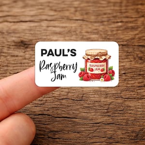 May include: A rectangular label with rounded corners, featuring the text "PAUL'S Raspberry Jam" in black. An illustration of a jam jar filled with red jam and raspberries is on the right. The label is held by a finger.