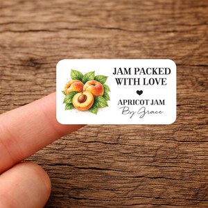 May include: A white label with rounded corners, displaying a watercolour illustration of apricots and green leaves. Text reads "JAM PACKED WITH LOVE" and "APRICOT JAM By Grace". The label is held by a finger.