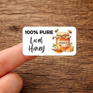 May include: A rectangular sticker with the words "100% Pure Local Honey" in black script. The sticker features a jar of honey with bees, flowers, and a honey dipper. The sticker is held by a finger.