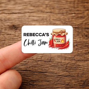 May include: A rectangular sticker with rounded corners. It features the text "REBECCA'S Chilli Jam" and an illustration of a jar of chilli jam with red chilli peppers. The jar has a label that says "Chilli Jam".
