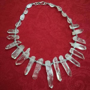 Puede incluir: Un collar de cristal de cuarzo transparente con un cierre plateado. El collar presenta cristales de cuarzo transparentes y puntiagudos y cuentas de cuarzo más pequeñas y redondeadas ensartadas en una cadena plateada. El fondo es rojo liso.
