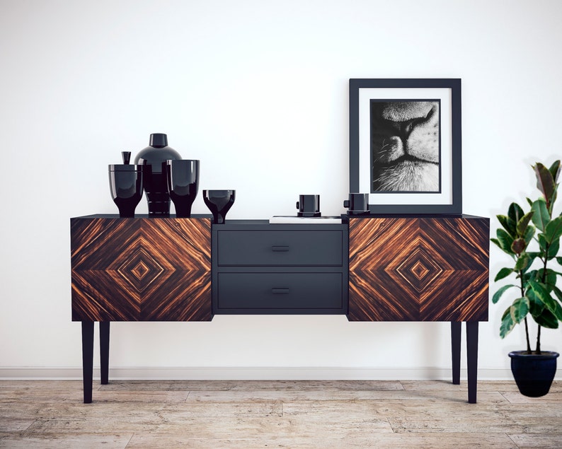May include: A black and brown wooden sideboard with two drawers and black legs. The sideboard is decorated with a geometric pattern. There are black vases and bowls on top of the sideboard. A black and white framed print of a cat's face is hanging on the wall above the sideboard. A potted plant is on the right side of the sideboard.