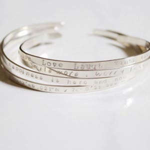 May include: Four silver bracelets stacked on top of each other. The bracelets are engraved with the words "Love Laugh", "Smile More", "Trust", "Worry Less", "Happiness is here", and "40.027°N / 74.0021°W".