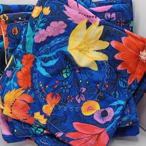 May include: Set of four blue fabric coasters with a floral pattern featuring red, yellow, pink, and orange flowers. The coasters have a white stitched border.