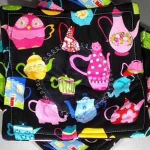 May include: Two black fabric pot holders with a white stitched border. The fabric features a repeating pattern of colorful teapots, owls, and houses.