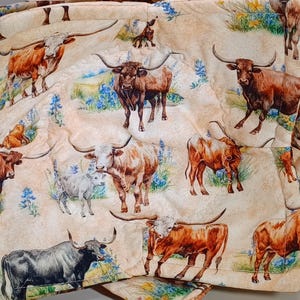 May include: A beige fabric with a repeating pattern of brown and white Texas longhorn cattle with blue wildflowers. The fabric is quilted and has a subtle texture.