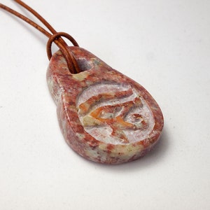 May include: A reddish-brown stone pendant with a carved Eye of Horus symbol, suspended from a brown leather cord. The pendant has a teardrop shape with a hole at the top. The carved symbol is in a lighter color, creating contrast.