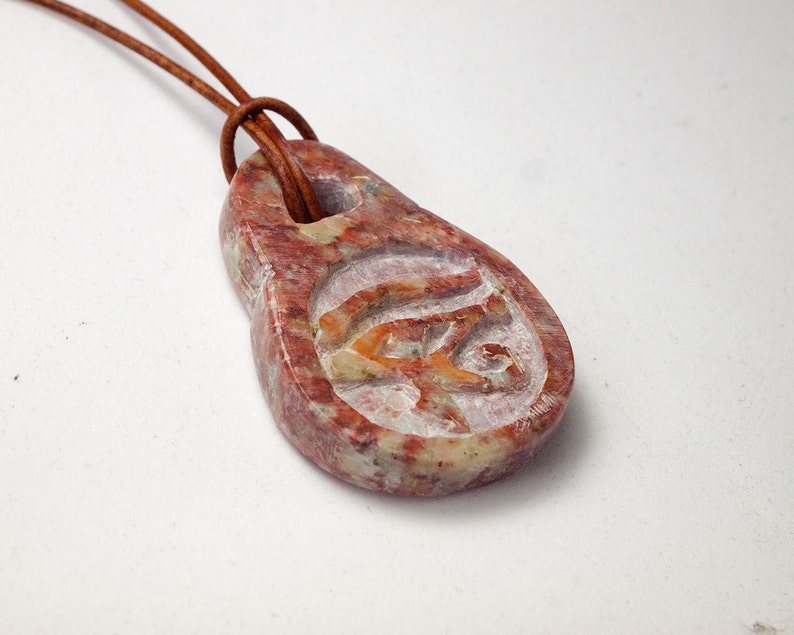 May include: A reddish-brown stone pendant with a carved Eye of Horus symbol, suspended from a brown leather cord. The pendant has a teardrop shape with a hole at the top. The carved symbol is in a lighter color, creating contrast.