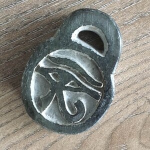 May include: A dark gray pendant with a carved Eye of Horus design. The pendant has a loop at the top and is set against a wooden surface. The Eye of Horus is a symbol of protection and royal power in ancient Egypt.
