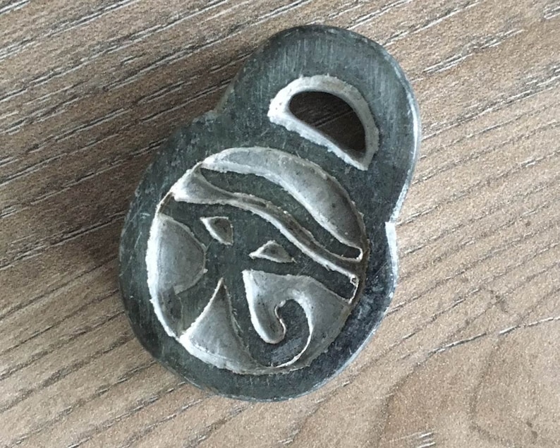 May include: A dark gray pendant with a carved Eye of Horus design. The pendant has a loop at the top and is set against a wooden surface. The Eye of Horus is a symbol of protection and royal power in ancient Egypt.
