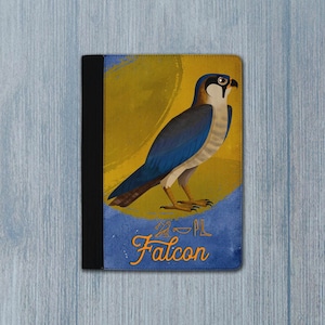 Könnte beinhalten: Ein Notizbuch mit einer Illustration eines blauen, weißen und beigen Falken. Der Falke ist vor einem gelben und blauen Hintergrund platziert. Das Wort "Falcon" ist in orangefarbener Schrift unter dem Vogel geschrieben. Das Notizbuch hat einen schwarzen Buchrücken.