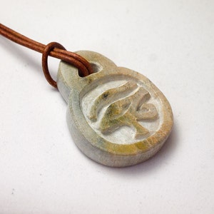 May include: A carved stone pendant with an Eye of Horus symbol, attached to a brown leather cord. The pendant is a light gray color with hints of green and yellow. The cord is knotted at the top.