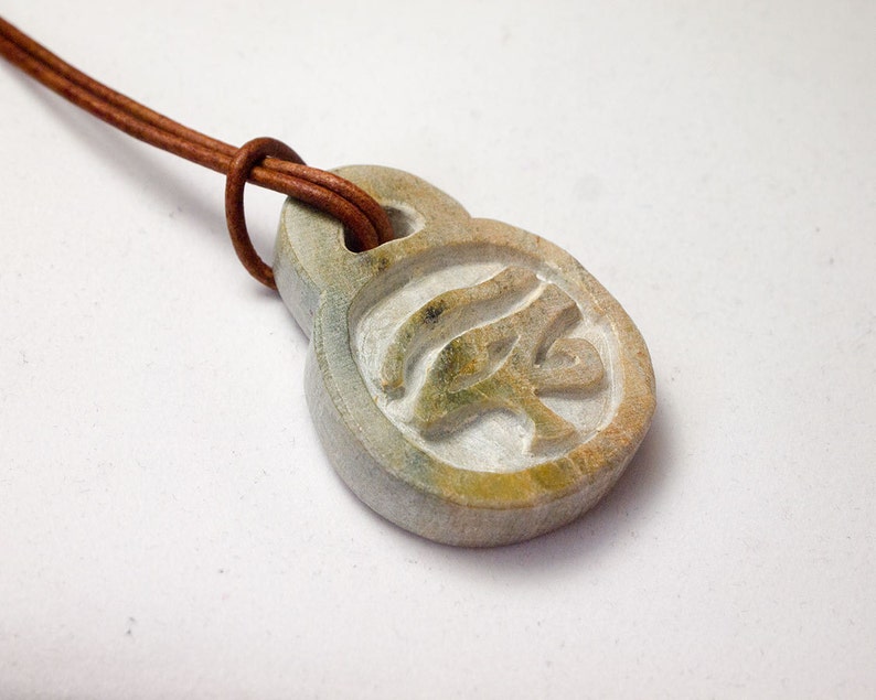 May include: A carved stone pendant with an Eye of Horus symbol, attached to a brown leather cord. The pendant is a light gray color with hints of green and yellow. The cord is knotted at the top.