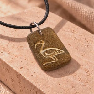 May include: A rectangular brown wooden pendant featuring an engraved ibis bird design. The pendant is suspended from a thin black cord by a silver-toned metal jump ring. The wood has a visible grain texture and rounded corners.