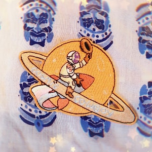 May include: A yellow and white embroidered patch of an astronaut riding a rocket with a planet ring around it. The text "Cosmic Cowboy" is embroidered on the ring.