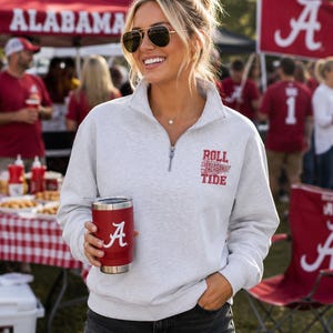 May include: A light gray quarter-zip pullover sweatshirt featuring a red embroidered pennant graphic with the text Roll Tide Alabama on the left chest. The garment is paired with dark gray denim shorts and a red insulated tumbler with a white A logo.