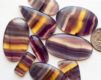 May include: Assortment of 10 colorful, striped, polished fluorite cabochons in shades of purple, yellow, and white. The cabochons are various shapes and sizes, with some being oval, teardrop, and rectangular.