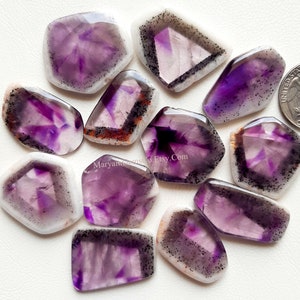 Puede incluir: Una colecci&oacute;n de 12 cabujones de amatista de diferentes formas y tama&ntilde;os. Los cabujones son una mezcla de morado y blanco con inclusiones negras.