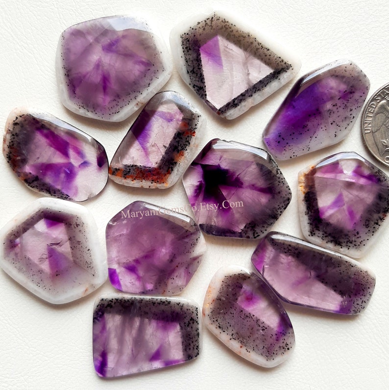 Puede incluir: Una colecci&oacute;n de 12 cabujones de amatista de diferentes formas y tama&ntilde;os. Los cabujones son una mezcla de morado y blanco con inclusiones negras.