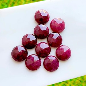 Puede incluir: Un conjunto de nueve rubíes redondos, facetados y de color rojo intenso dispuestos en un grupo. Cada piedra preciosa presenta un patrón simétrico de talla rosa con múltiples facetas triangulares que reflejan la luz, mostrando un tono carmesí oscuro uniforme.