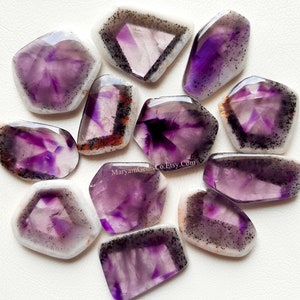 Puede incluir: Una colecci&oacute;n de 11 cabujones de amatista p&uacute;rpura con un borde blanco y negro. Las piedras tienen una forma irregular y un aspecto natural y en bruto.