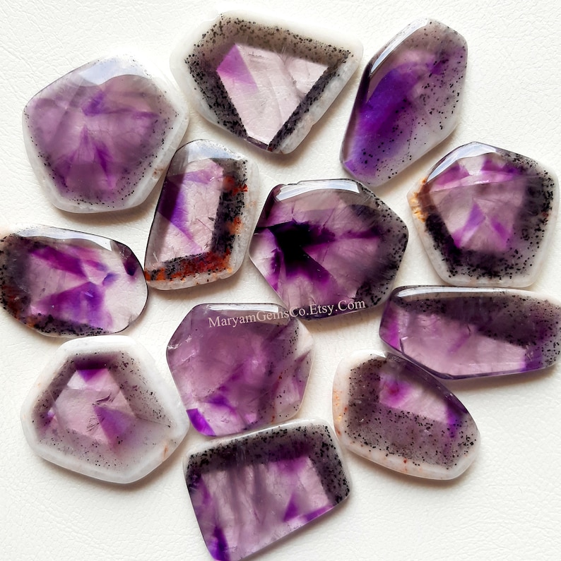 Puede incluir: Una colecci&oacute;n de 11 cabujones de amatista p&uacute;rpura con un borde blanco y negro. Las piedras tienen una forma irregular y un aspecto natural y en bruto.