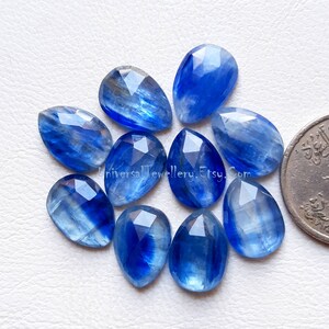 May include: Ten blue kyanite pear-shaped gemstones, each with a unique striated pattern. The stones are approximately 15mm in length and are arranged in a circular pattern on a white surface.