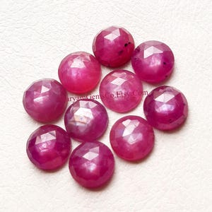 May include: Nine faceted, round ruby gemstones in a deep pinkish-red hue. The gems have a polished, sparkling appearance. The gemstones are displayed on a white surface. Each gem is approximately 1 cm in diameter.