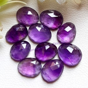 May include: A collection of ten faceted amethyst gemstones. The gemstones are oval-shaped with a deep purple hue, and each stone has a polished, reflective surface. The gemstones are arranged on a white surface.
