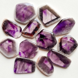 Puede incluir: Una colecci&oacute;n de 12 cabujones de amatista, cada uno con una variedad de tonos morados e inclusiones blancas y negras. Los cabujones tienen una forma irregular y un acabado liso y pulido.