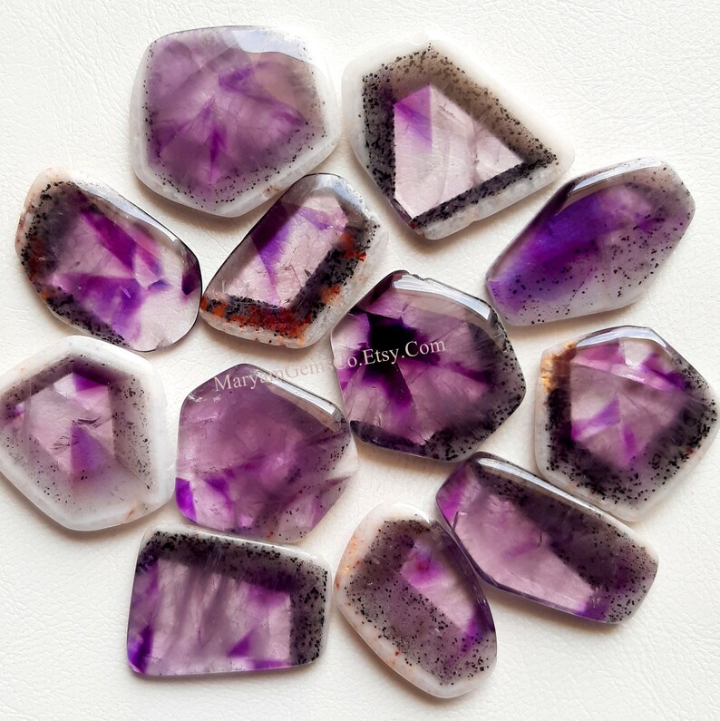 Puede incluir: Una colecci&oacute;n de 12 cabujones de amatista, cada uno con una variedad de tonos morados e inclusiones blancas y negras. Los cabujones tienen una forma irregular y un acabado liso y pulido.