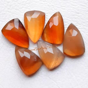 May include: Six faceted shield-shaped carnelian gemstones in varying shades of orange and amber. The stones feature a polished surface with geometric cuts. The text "VersaJewellery.Etsy.com" is printed in small brown letters across the center.