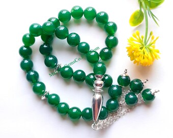 Può includere: Una collana di perline verdi con un ciondolo e una catena d'argento. Le perline sono rotonde e lucenti. Un piccolo fiore giallo e foglie verdi sono sullo sfondo. Il testo "MarGemsCo.Com" è visibile sulla collana.