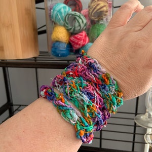 May include: A colourful crocheted bracelet made from multiple strands of yarn in shades of pink, purple, blue, green, orange, and red.