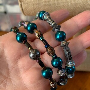May include: Three teal beaded bracelets with decorative metal elements. The bracelets feature a mix of round and cylindrical beads, with silver and copper-toned metal accents. The bracelets are designed to wrap around the wrist.